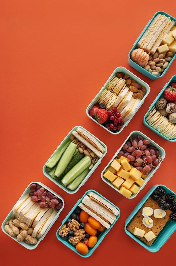 Flat lay of assorted healthy lunchboxes with fruits and nuts against vibrant orange background.